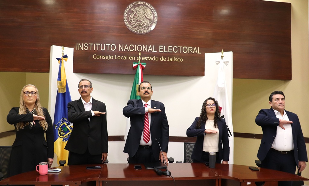 tres hombres y dos mujeres de pie con la mano derecha en el pecho, mirando hacia el frente