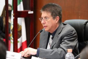 hombre de lentes y traje gris, sentado frente a un micrófono la toma es lateral
