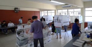 varias personas al interior de un salón de clases que tiene urnas y mamparas del INE