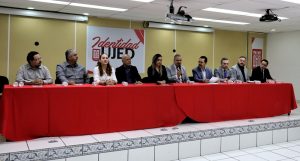 Dos mujeres y ocho hombres sentados frente a una mesa con mantel rojo y micrófonos