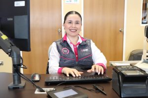 Mujer cabello recogido y con uniforme gris con rosa sentada viendo a la cámara
