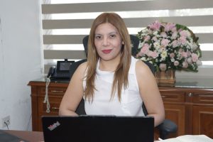 Mujer cabello castaño claro sentada frente a una computadora negra, vestida de blanco.
